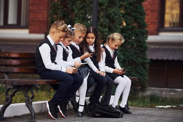 Skolbarn Uniform Som Sitter Utomhus Bänken Med Smartphones — Stockfoto