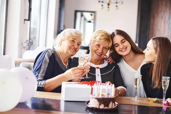 Glasses with alcohol in hands and cake on table. Senior woman with family and friends celebrating a birthday indoors.