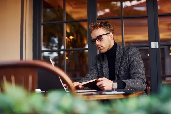 エレガントな若いです男でフォーマル摩耗座っていますカフェで彼のラップトップと本を読みます — ストック写真