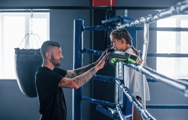 stock image Conception of assistance. Young tattooed boxing coach teaches cute little girl in the gym.