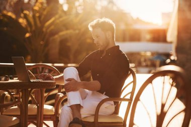 Masanın yanında oturuyor. Genç adam güneşli bir günde dışarıda. Tatil kavramı.