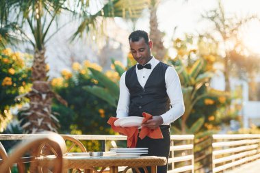 Tabakları kırmızı bezle siler. Resmi kıyafetli zenci garson güneşli bir günde iş yerinde..