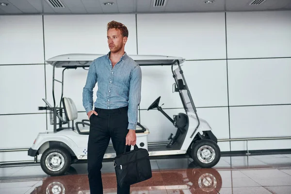 Against vehicle. Young man in formal wear is in modern airport. Conception of vacation.