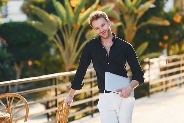 Dizüstü bilgisayarı var. Genç adam güneşli bir günde dışarıda. Tatil kavramı.