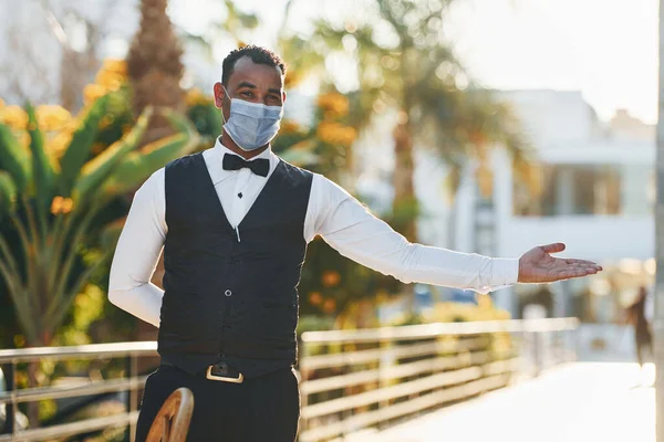 In protective mask. Black waiter in formal clothes is at his work outdoors at sunny daytime.