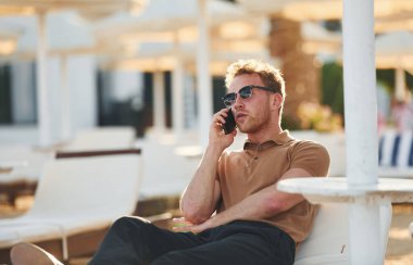 Oturuyor ve telefon kullanıyor. Genç adam güneşli bir günde dışarıda. Tatil kavramı.