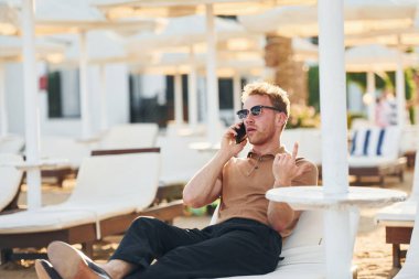 Oturuyor ve telefon kullanıyor. Genç adam güneşli bir günde dışarıda. Tatil kavramı.