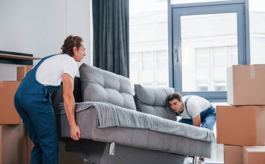 Ağır kanepe taşıyor. İçeride çalışan mavi üniformalı iki genç nakliyeci..