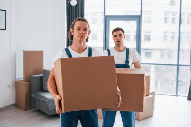 Ellerinde kutularla. İçeride çalışan mavi üniformalı iki genç nakliyeci..