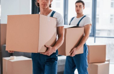 Ellerinde kutularla. İçeride çalışan mavi üniformalı iki genç nakliyeci..