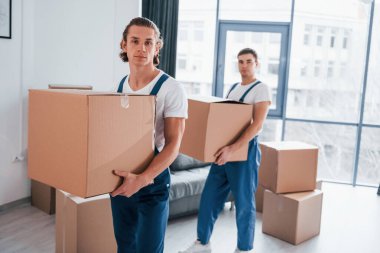 Ellerinde kutularla. İçeride çalışan mavi üniformalı iki genç nakliyeci..