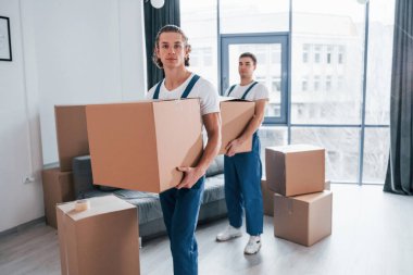 Ellerinde kutularla. İçeride çalışan mavi üniformalı iki genç nakliyeci..