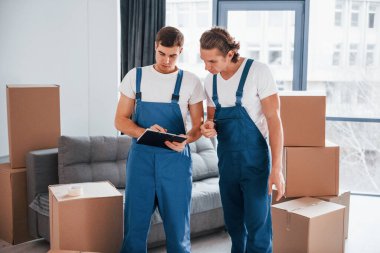 İçinde belge olan bir not defteri tutuyor. İçeride çalışan mavi üniformalı iki genç nakliyeci..