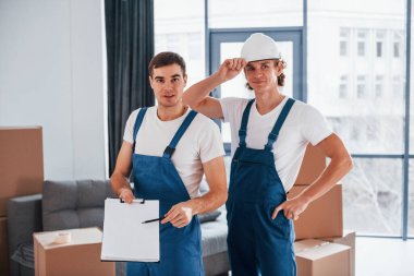 Document to sign. Two young movers in blue uniform working indoors in the room.