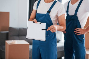 Holds notepad with document. Two young movers in blue uniform working indoors in the room.