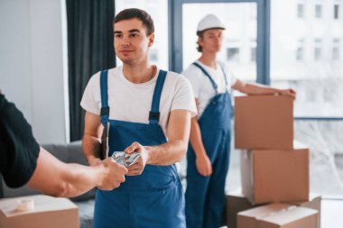 Man takes money from another guy. Two young movers in blue uniform working indoors in the room.