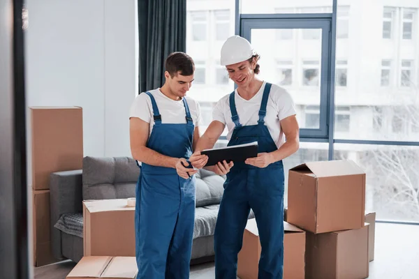 Holds notepad with document. Two young movers in blue uniform working indoors in the room.