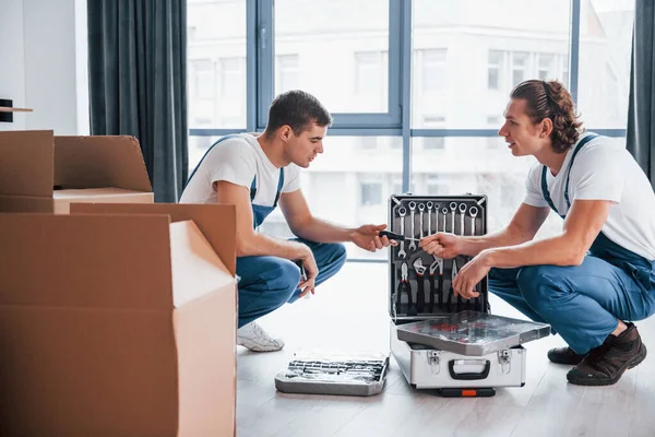 Uses tools from case. Two young movers in blue uniform working indoors in the room.