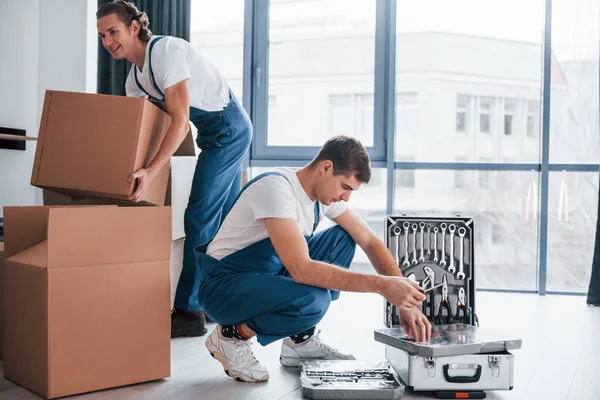 Uses tools from case. Two young movers in blue uniform working indoors in the room.