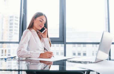 Telefonla konuş. Beyaz elbiseli genç kadın modern ofiste kapalı bir yerde..