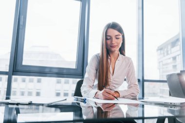 Masanın yanında oturuyor. Beyaz elbiseli genç kadın modern ofiste kapalı bir yerde..