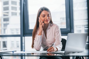 Have conversation by the phone. Young woman in white formal clothes is indoors in the modern office.