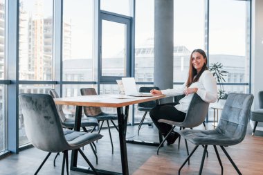 Masanın yanında oturuyor. Beyaz elbiseli genç kadın modern ofiste kapalı bir yerde..