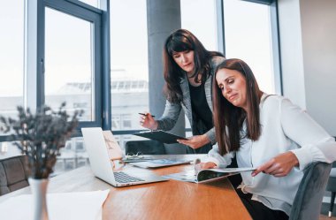 Two women in formal clothes is indoors in the modern office works together.