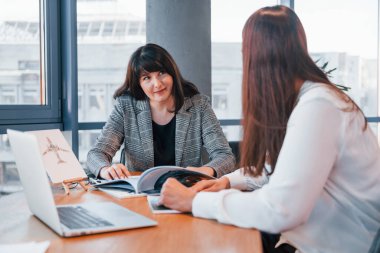 Masanın yanında oturuyor. Modern ofiste resmi giyinmiş iki kadın birlikte çalışıyor..