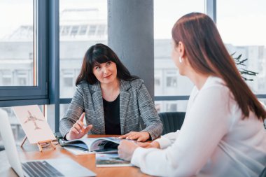 Masanın yanında oturuyor. Modern ofiste resmi giyinmiş iki kadın birlikte çalışıyor..