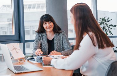 Masanın yanında oturuyor. Modern ofiste resmi giyinmiş iki kadın birlikte çalışıyor..