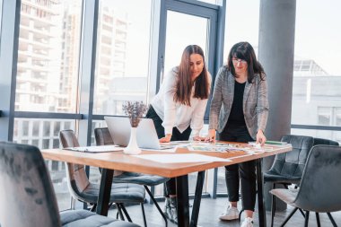 Ayakta dikilip konuşuyorum. Modern ofiste resmi giyinmiş iki kadın birlikte çalışıyor..