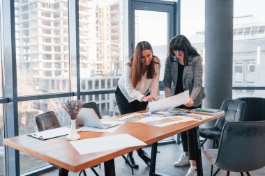 Ayakta dikilip konuşuyorum. Modern ofiste resmi giyinmiş iki kadın birlikte çalışıyor..