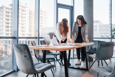 Ayakta dikilip konuşuyorum. Modern ofiste resmi giyinmiş iki kadın birlikte çalışıyor..
