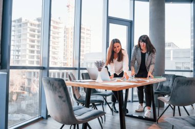Ayakta dikilip konuşuyorum. Modern ofiste resmi giyinmiş iki kadın birlikte çalışıyor..