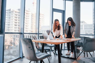 Ayakta dikilip konuşuyorum. Modern ofiste resmi giyinmiş iki kadın birlikte çalışıyor..