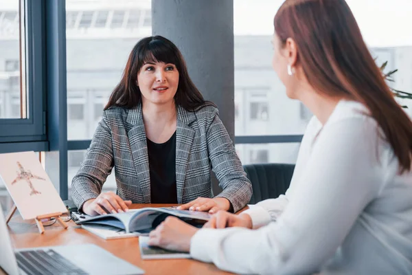 Masanın yanında oturuyor. Modern ofiste resmi giyinmiş iki kadın birlikte çalışıyor..