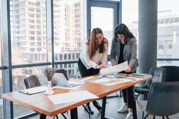 Ayakta dikilip konuşuyorum. Modern ofiste resmi giyinmiş iki kadın birlikte çalışıyor..