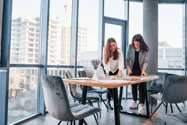 Ayakta dikilip konuşuyorum. Modern ofiste resmi giyinmiş iki kadın birlikte çalışıyor..