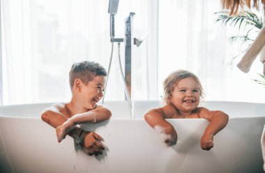 Two kids having fun and washing themselves in the bath at home. Posing for a camera.