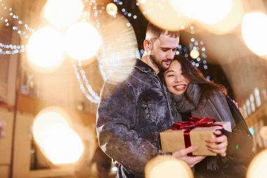 Holds gift box. Happy multiracial couple together outdoors in the city celebrating New year.