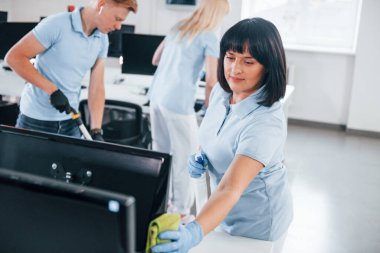 Cleans monitor. Group of workers clean modern office together at daytime.