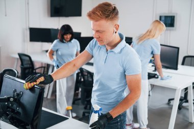 Cleans monitor. Group of workers clean modern office together at daytime.