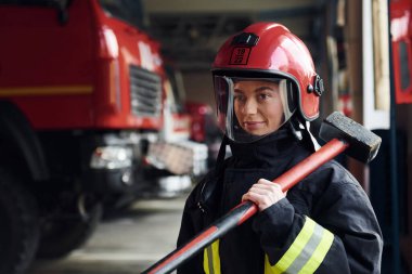 With hammer in hands. Female firefighter in protective uniform standing near truck.