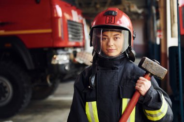 With hammer in hands. Female firefighter in protective uniform standing near truck.