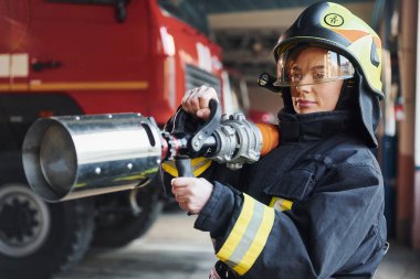 Hose in hands. Female firefighter in protective uniform standing near truck.