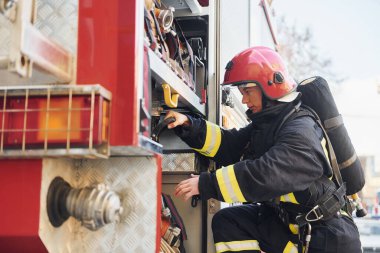 Takes equipment. Female firefighter in protective uniform standing near truck.