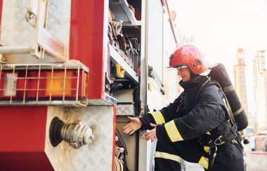 Takes equipment. Female firefighter in protective uniform standing near truck.