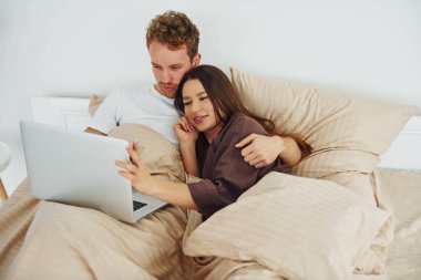 Çift dizüstü bilgisayarla yatakta yatıyor. Gündüz vakti güzel, modern yatak odasının iç tasarımı ve tasarımı..