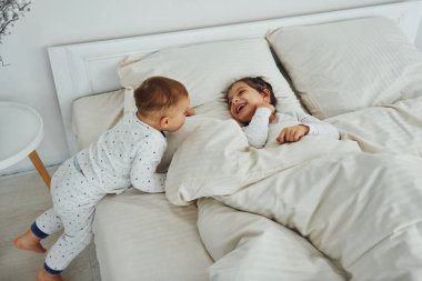 Two kids have fun while liyng down on bed. Interior and design of beautiful modern bedroom at daytime.